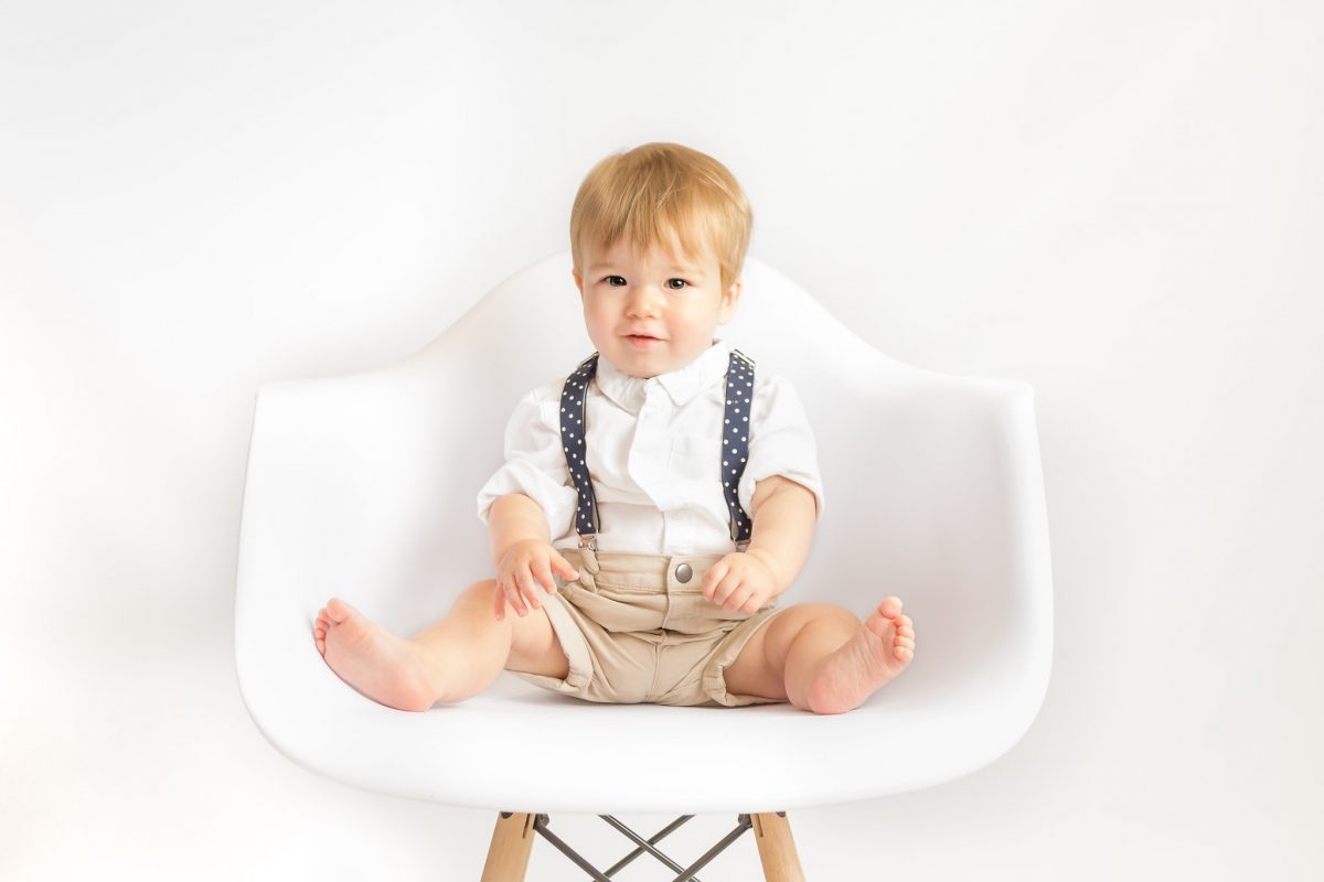 A baby sits in a white chair, looking in at the camera for his photoshoot
