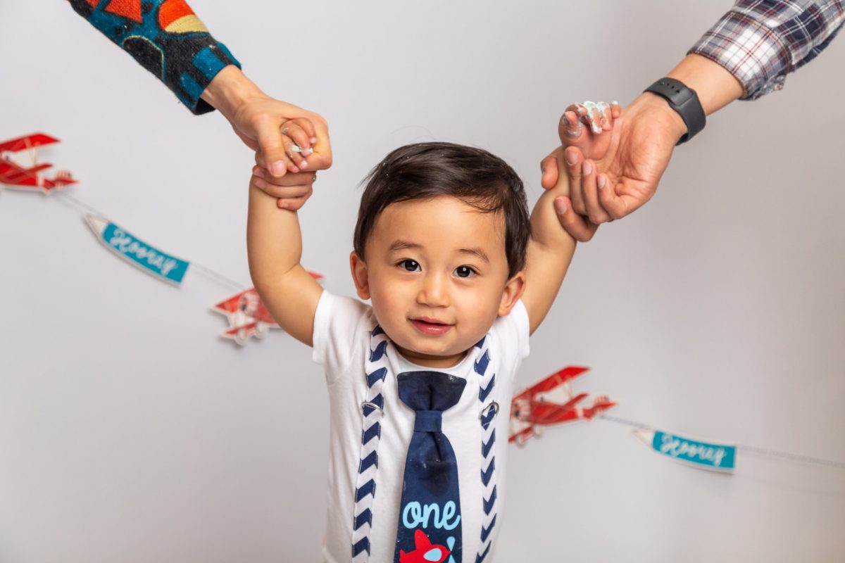 A baby smiles at the camera during his first birthday photoshoot. He is stood, and holding hands with his parents, just the parents hands are in the photo with the baby