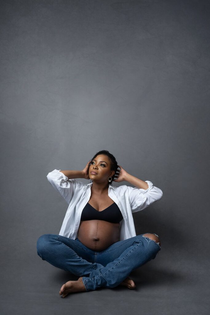 Pregnant woman sitting cross-legged in white shirt and jeans with hands in hair, casual maternity portrait on grey background