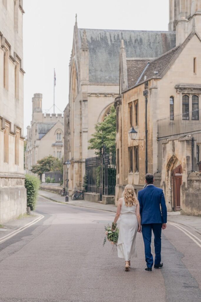 Bride and groom walking hand in hand down historic Oxford street near Oxford Town hall. The street is lined with Gothic architecture and church spires