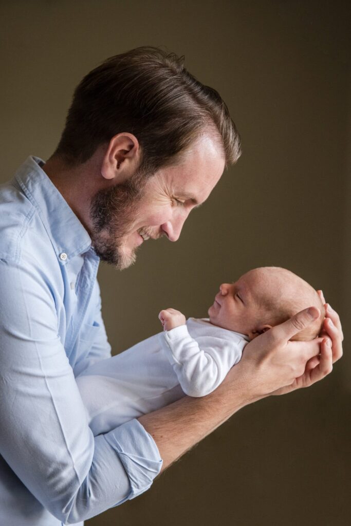 Father and newborn baby smile at each other as he cradles the baby in his arms.