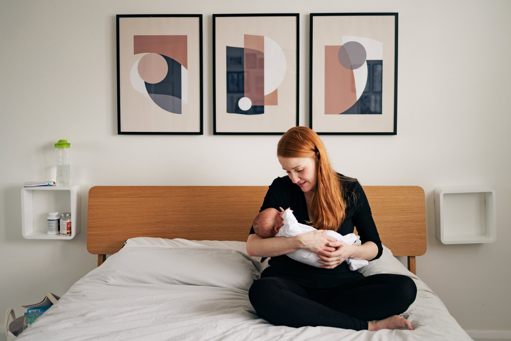 A photograph capturing a new mum cradling her baby sat on her bed at home