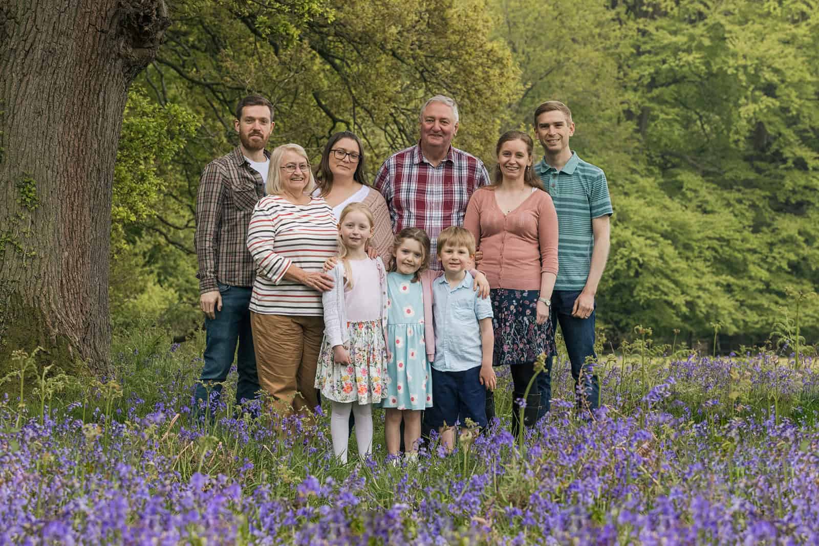 An extended family with 3 generations stand together for a photo with bluebells in the foreground in the Ashridge Estate