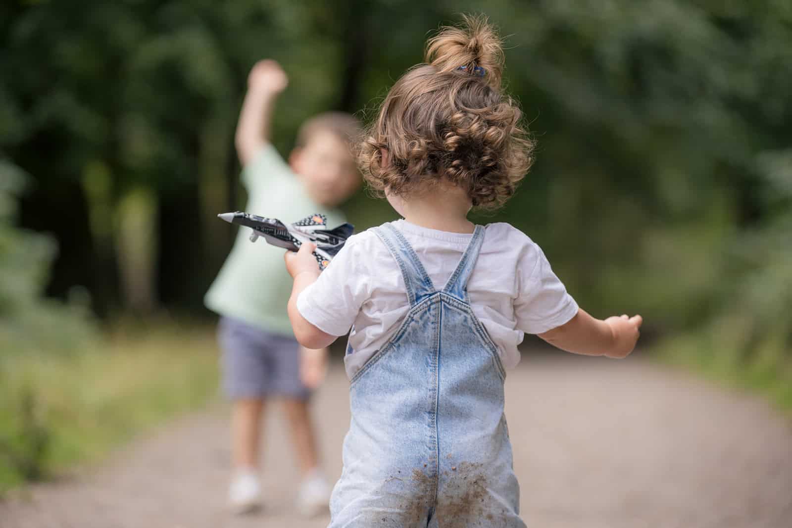 A toddler flies a plane in her hand while running away from her camera, her brother can be seem in the distance taking a similar action towards the camera