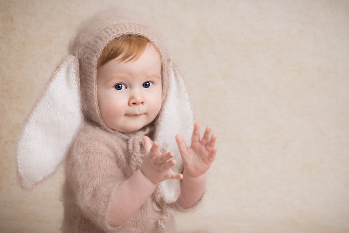 A toddler is dressed in a soft easter bunny outfit, and claps their hands while smiling at the camera during a photoshoot in Berkhamsted.