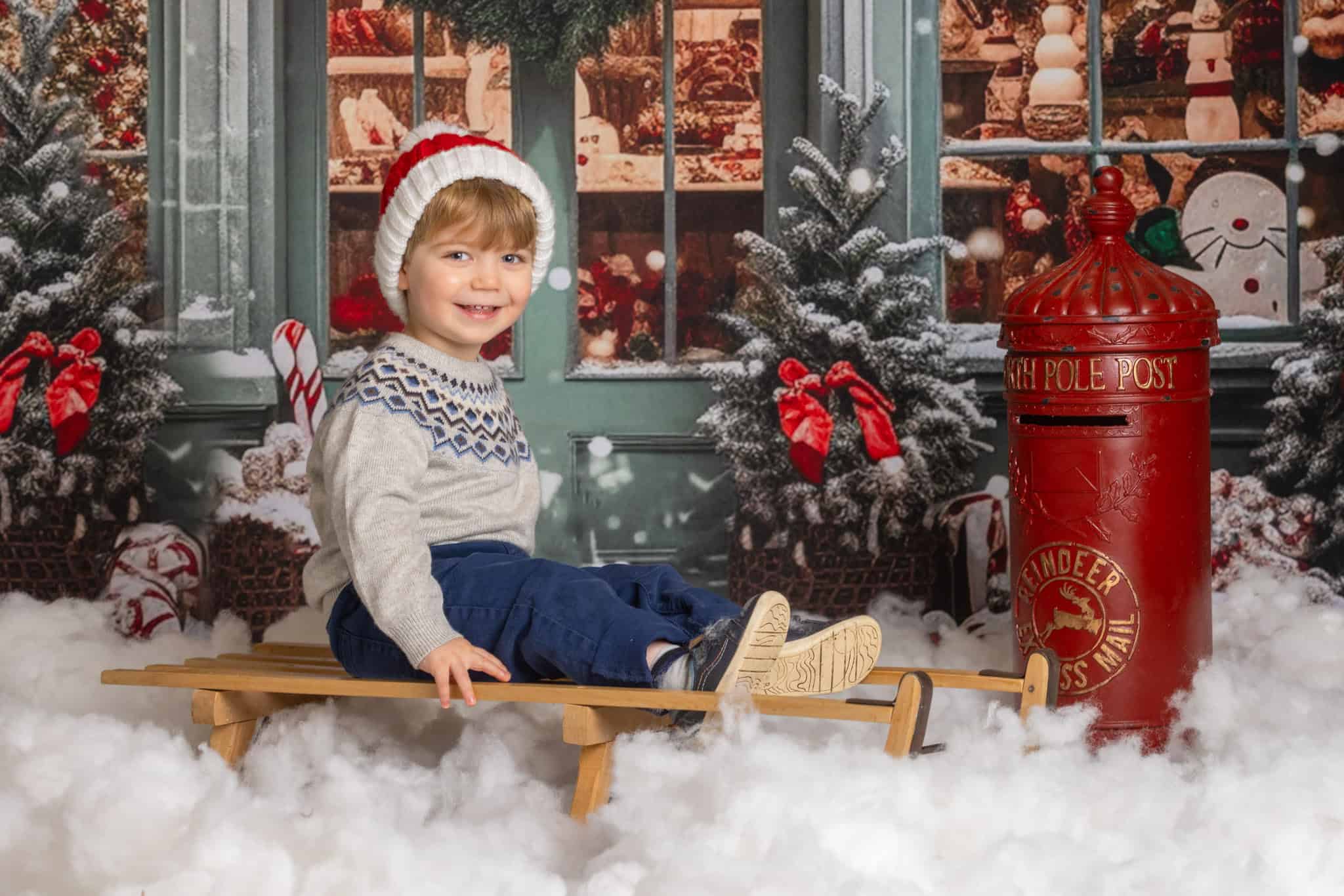 A boy sits on a sledge, surrounded by snow and next to a festive red postbox during an indoor Christmas photoshoot at the Ashridge Estate