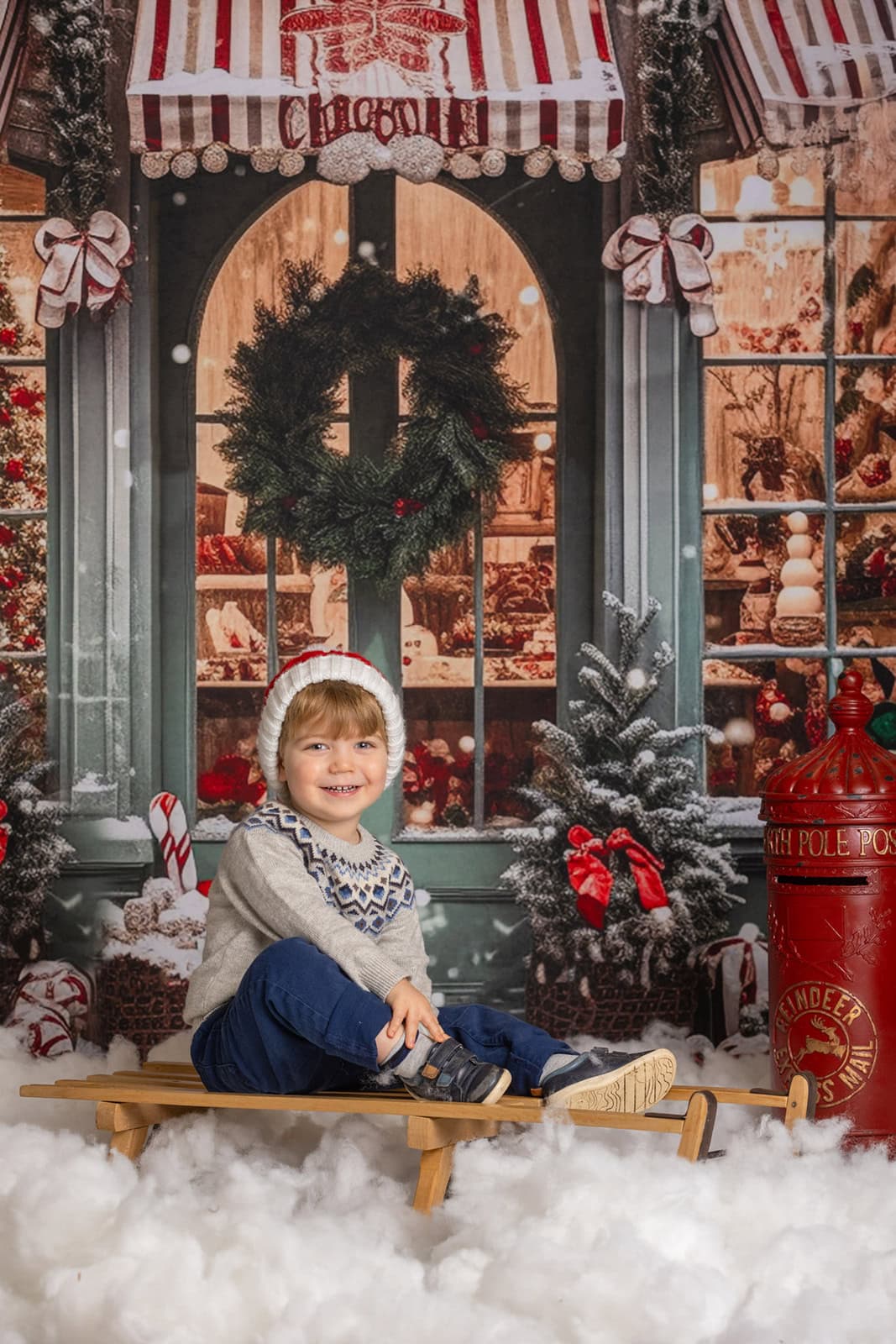 Boy sits on a wooden sledge and smiles at the camera during a Christmas photoshoot at Ashridge. There's a red Christmas postbox, lots of pretend snow and a snowy street scene.